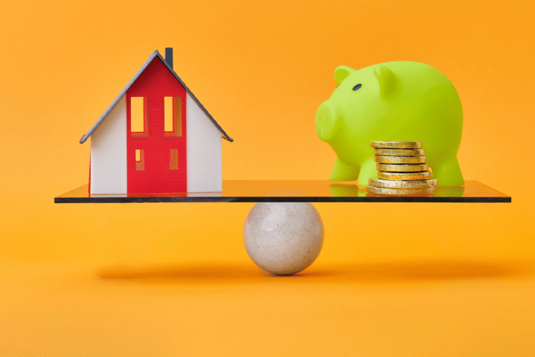 Scale balances a small house model against a green piggy bank with coins on the other side, against an orange backdrop; conveys housing versus savings.