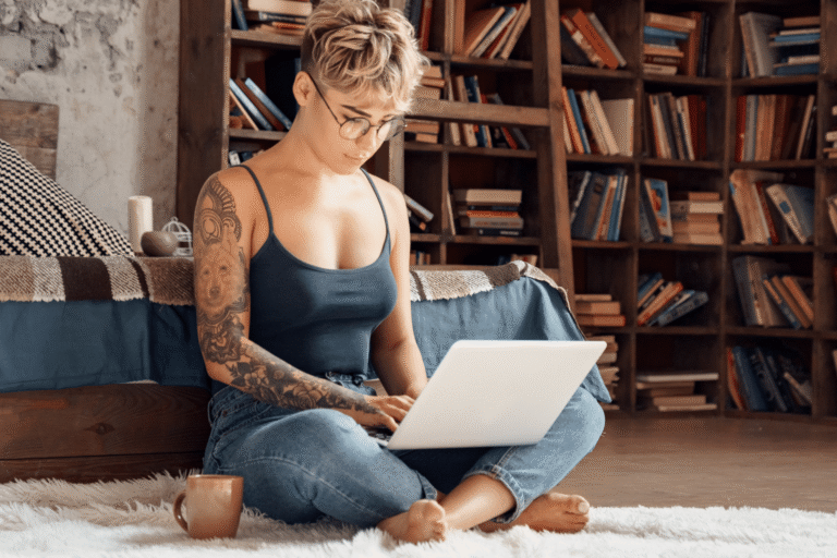 Individual with short blond hair and glasses sits cross-legged on a white rug, typing on a laptop with a book-filled wooden bookshelf behind.