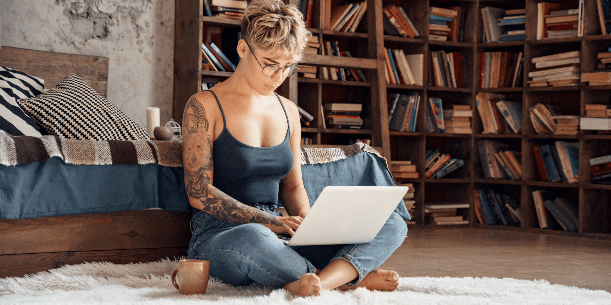 LJHHL_Blog_Side_Hustles Individual with short blond hair and glasses sits cross-legged on a white rug, typing on a laptop with a book-filled wooden bookshelf behind.