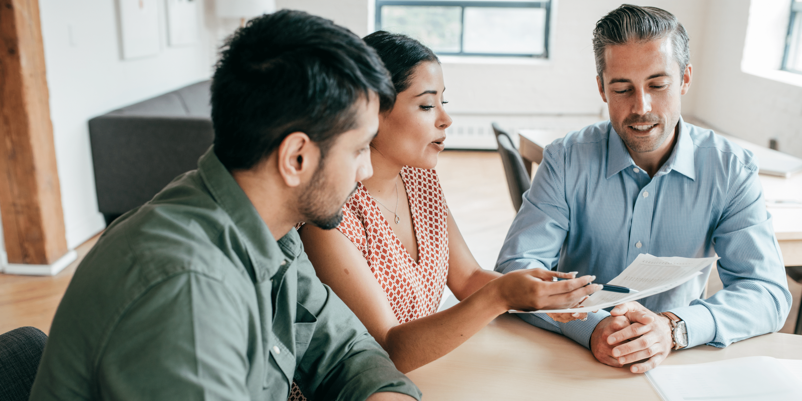 Three coworkers in a bright office review documents together at a round table, collaborating on a project.