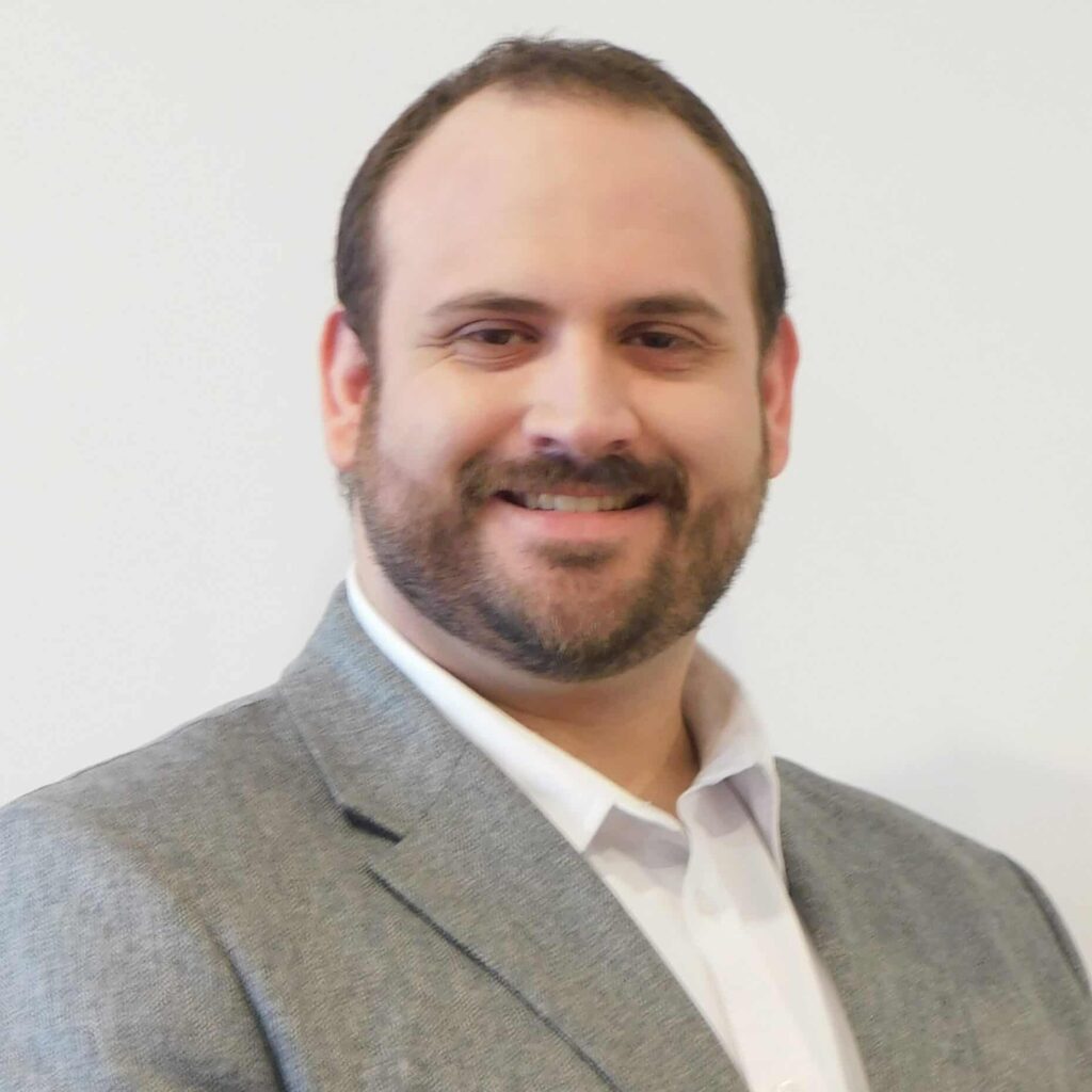 VOELKEL Tallen - Profile Image Smiling man wearing a light gray blazer and white shirt, head and shoulders visible against a neutral background.