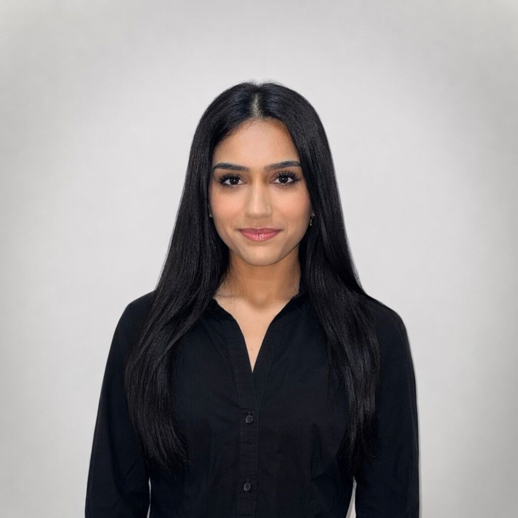 Asmi Soodan - Photo Head-and-shoulders portrait of a woman with long black hair, wearing a black blouse, against a light gray studio background with a subtle smile.